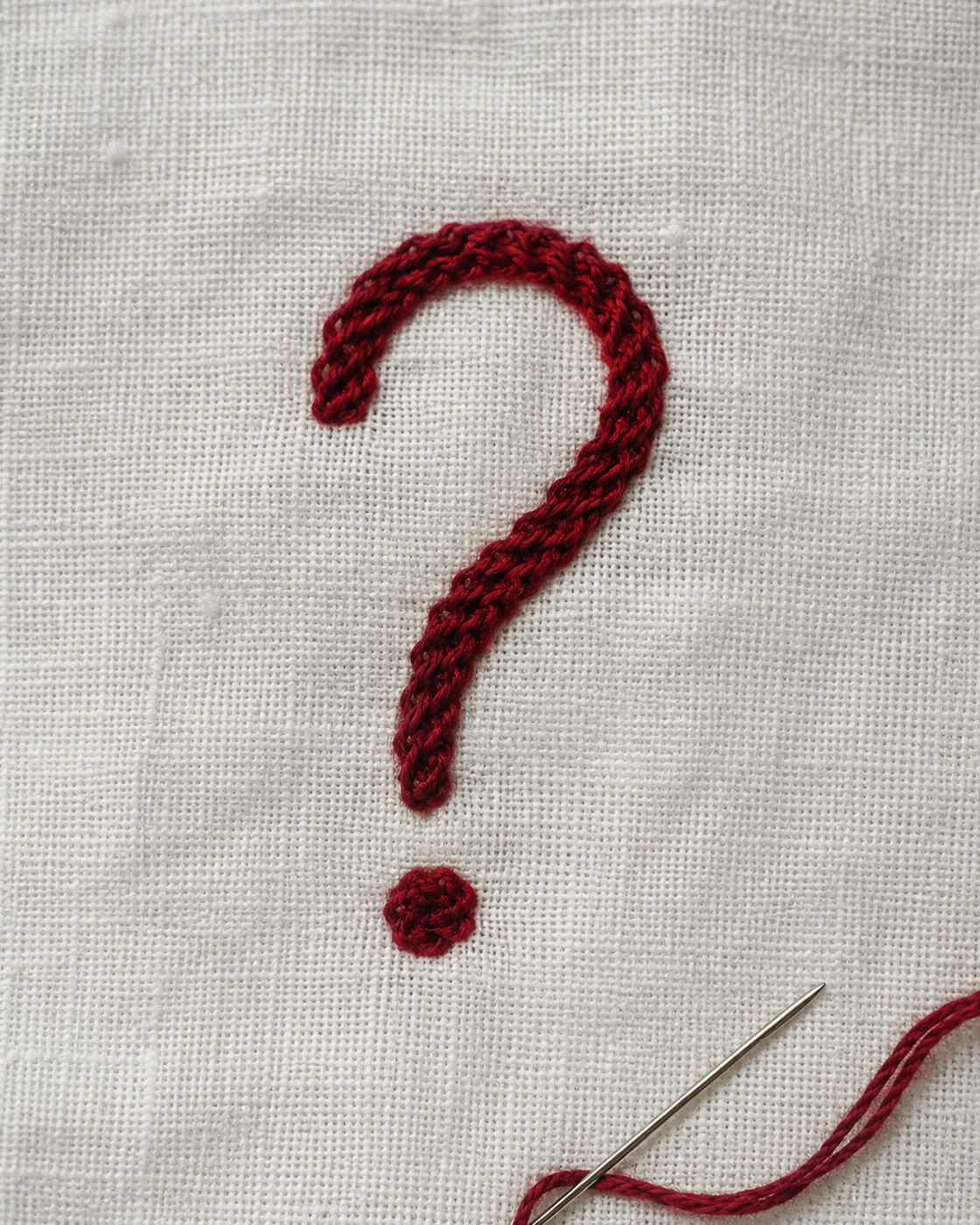 A red question mark embroidered on white linen with a needle